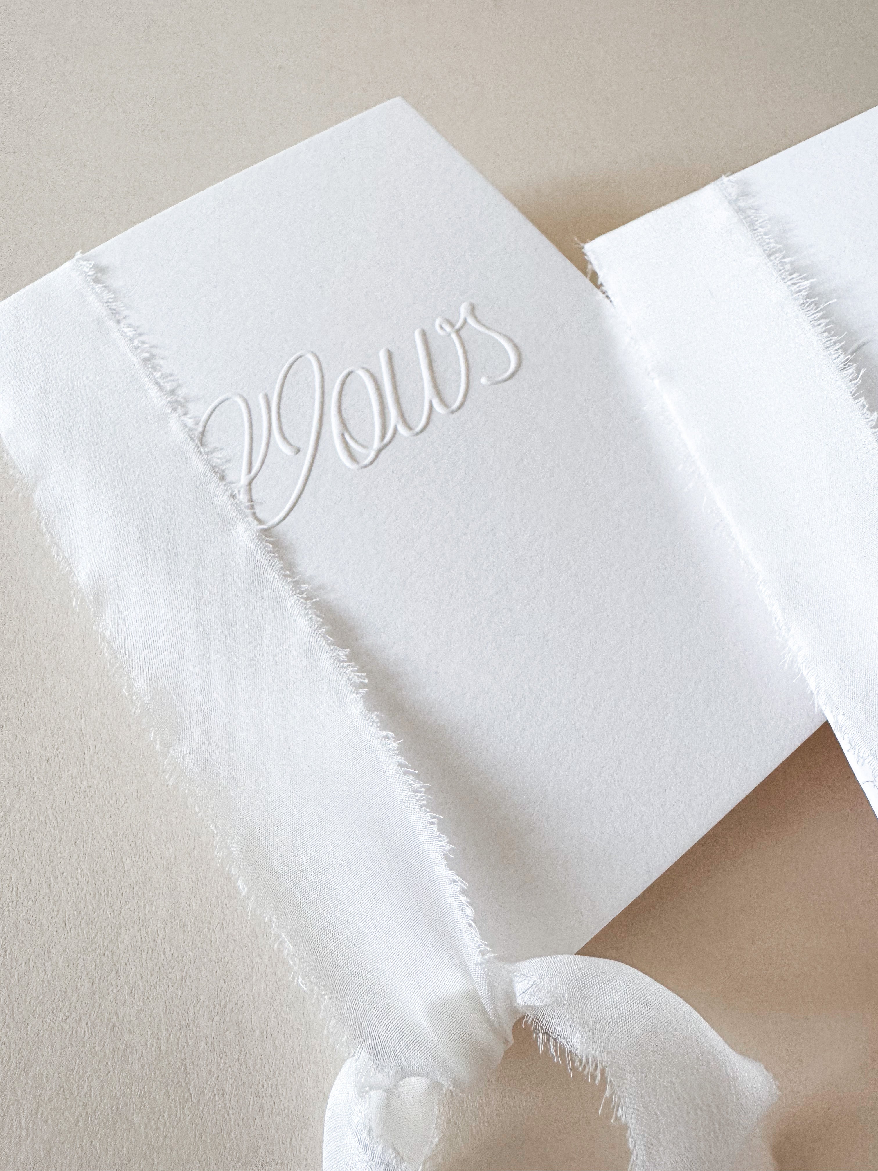 Handmade wedding Vows Book, made from white cotton card, with the text 'Vows' embossed on the front cover, tied together with white silk ribbon.