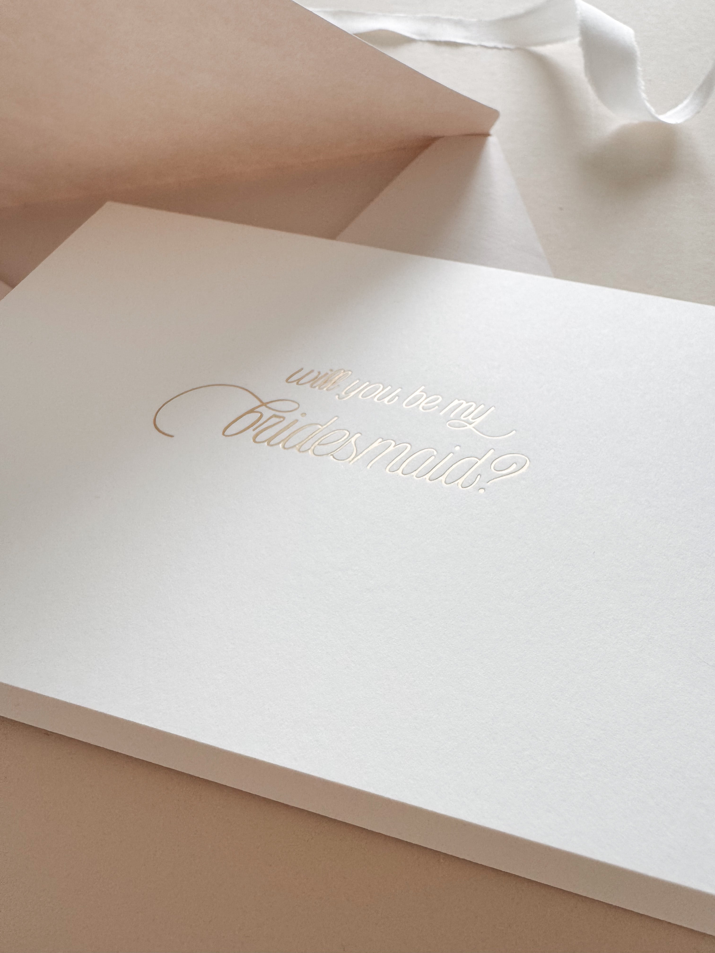 A hand-stamped bridesmaid proposal card with gold foil on a warm white cardstock surface, the text reads 'Will you be my Bridesmaid?'. A premium Almond coloured envelope is partially visible.
